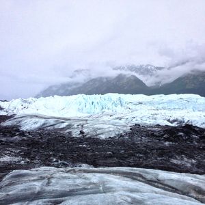 A serrated glacial wall, with dirty, gritty ice in the foreground and cloud covered mountains in the background. The photo almost appears black and white, aside from the teal blue of the seracs A serrated glacial wall, with dirty, gritty ice in the foreground and cloud covered mountains in the background. The photo almost appears black and white, aside from the teal blue of the seracs